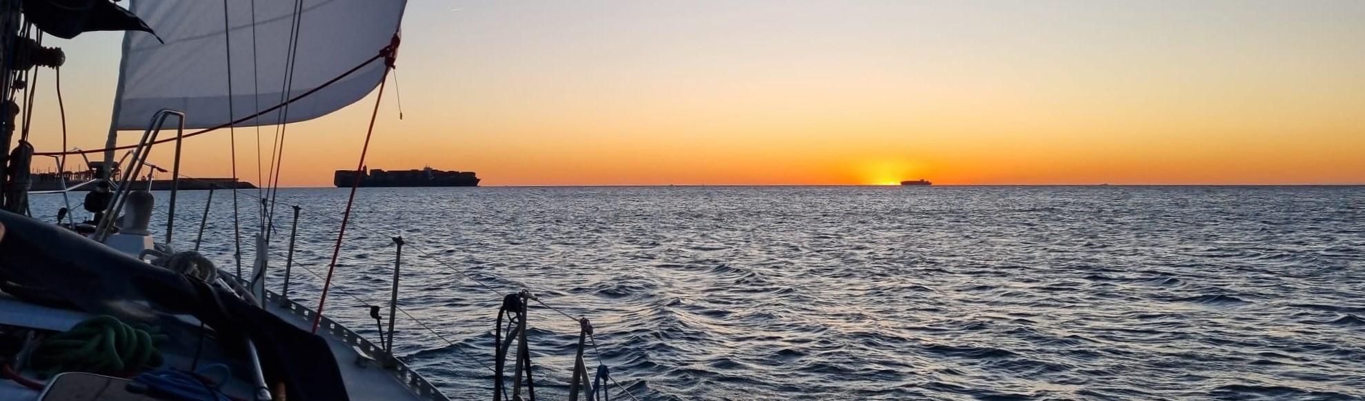 Desde un velero sale el sol al lado de un barco y a la izquierda un portacontenedores. Curso PER
